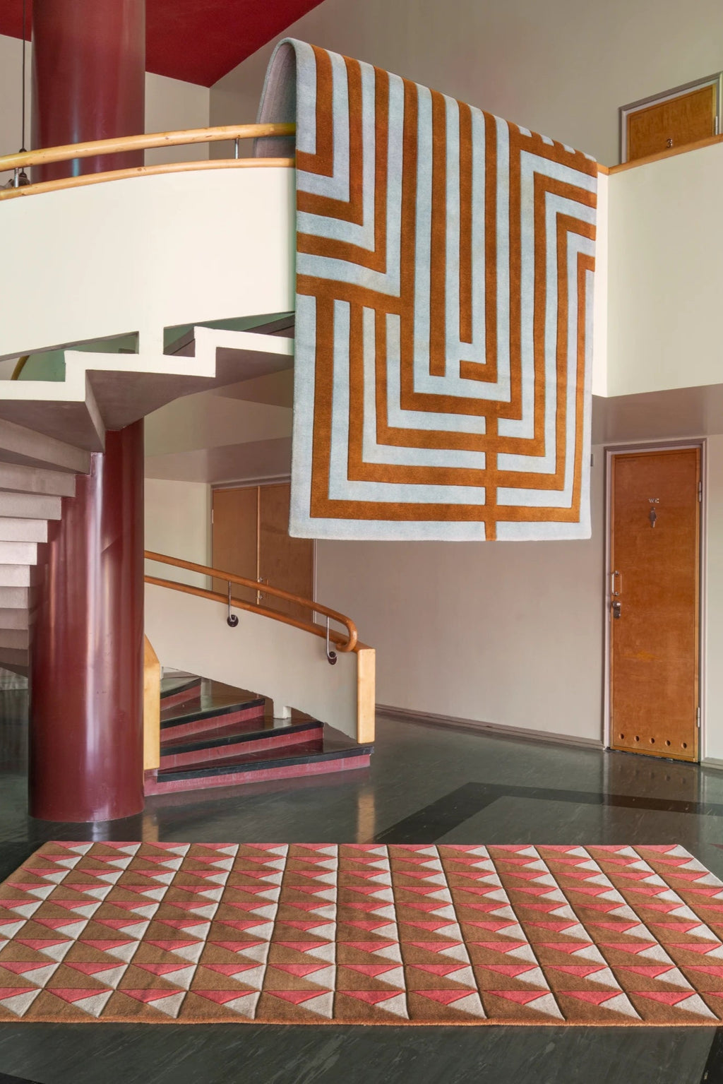 Layered Letters Wool Rug, featuring a geometric pattern in brown and light blue, draped over a banister, and a matching brown, red, and white patterned rug on the floor. Perfect for adding modern,...