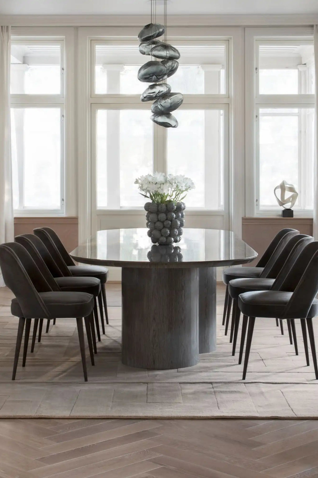 Layered Byzantine Grand Wool Rug, a large, light beige hand-tufted wool rug with subtle geometric patterns, centered under a modern dining table. Perfect for adding warmth and texture to contempora...