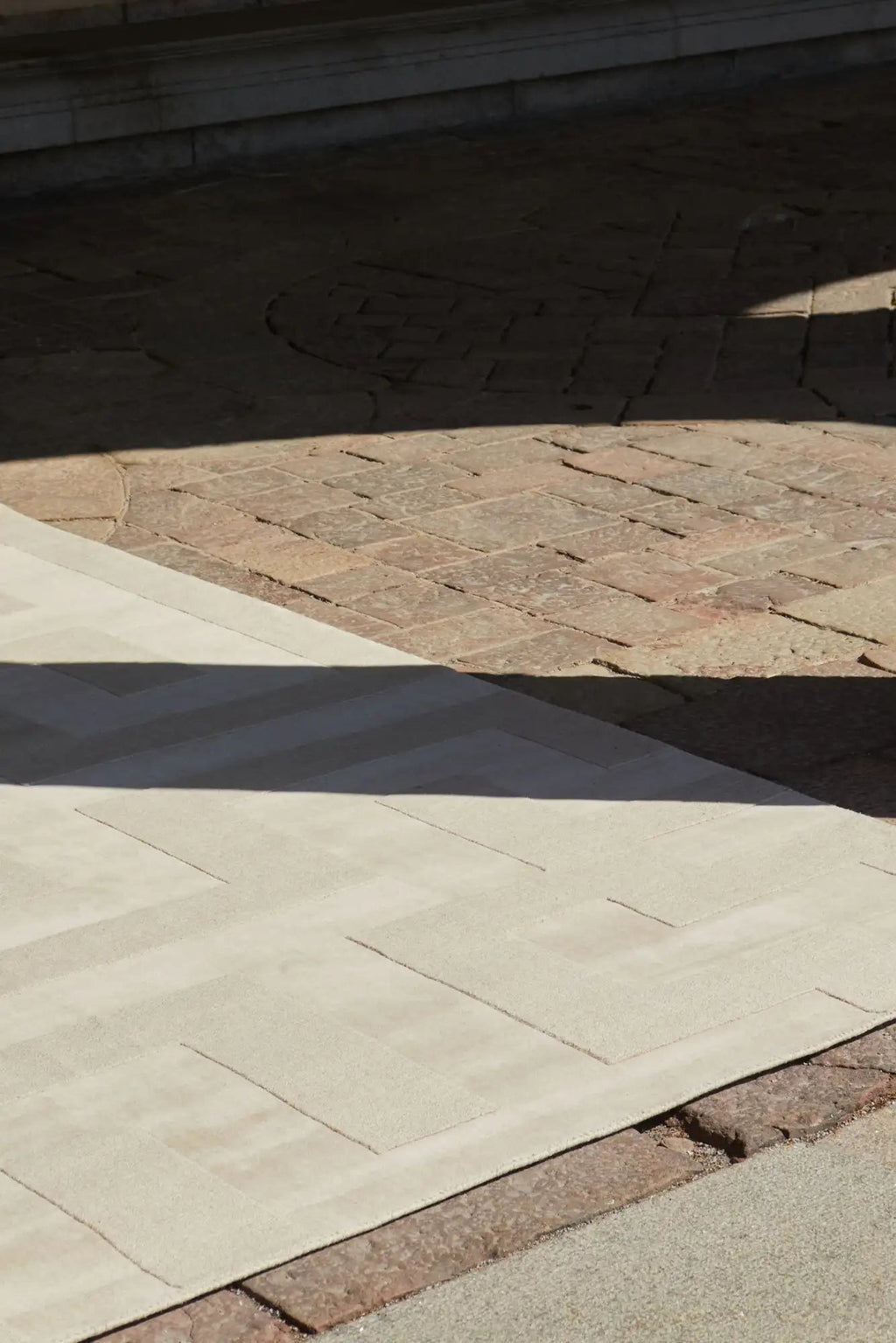 Layered Byzantine Grand Wool Rug, a light beige wool area rug with a subtle geometric pattern, partially illuminated by sunlight and partially in shadow, laid over a rustic brick patio. Perfect for...