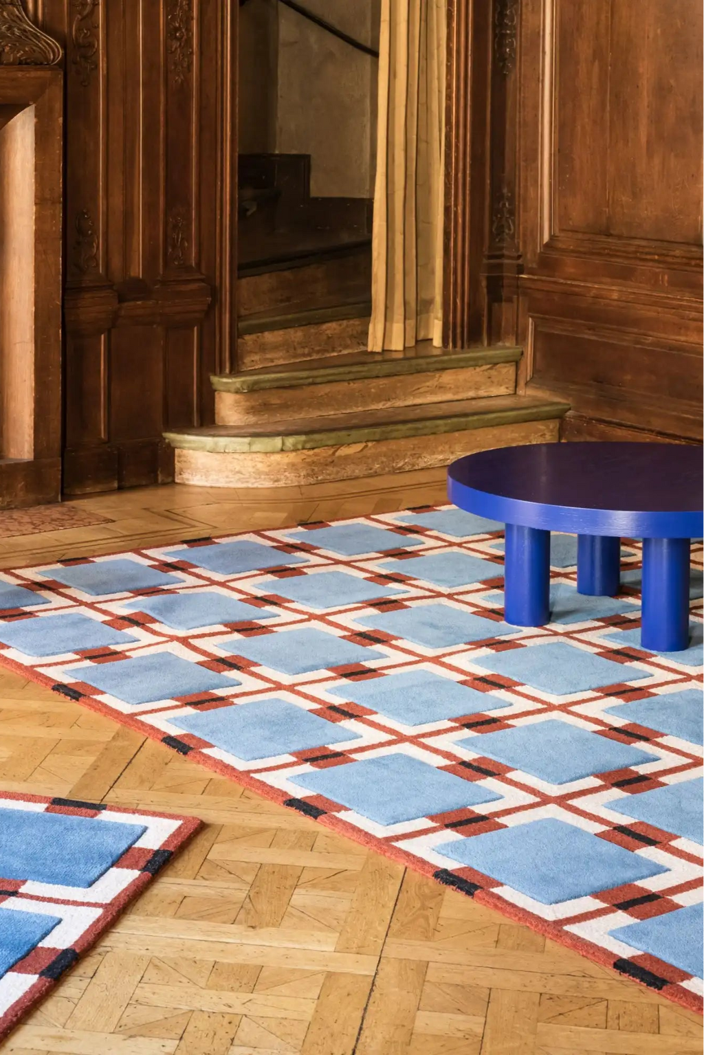 Layered Berry Rain Wool Rug, a blue, white, and terracotta geometric patterned carpet with black accents, complementing a blue coffee table on a hardwood floor. Perfect for adding a modern statemen...