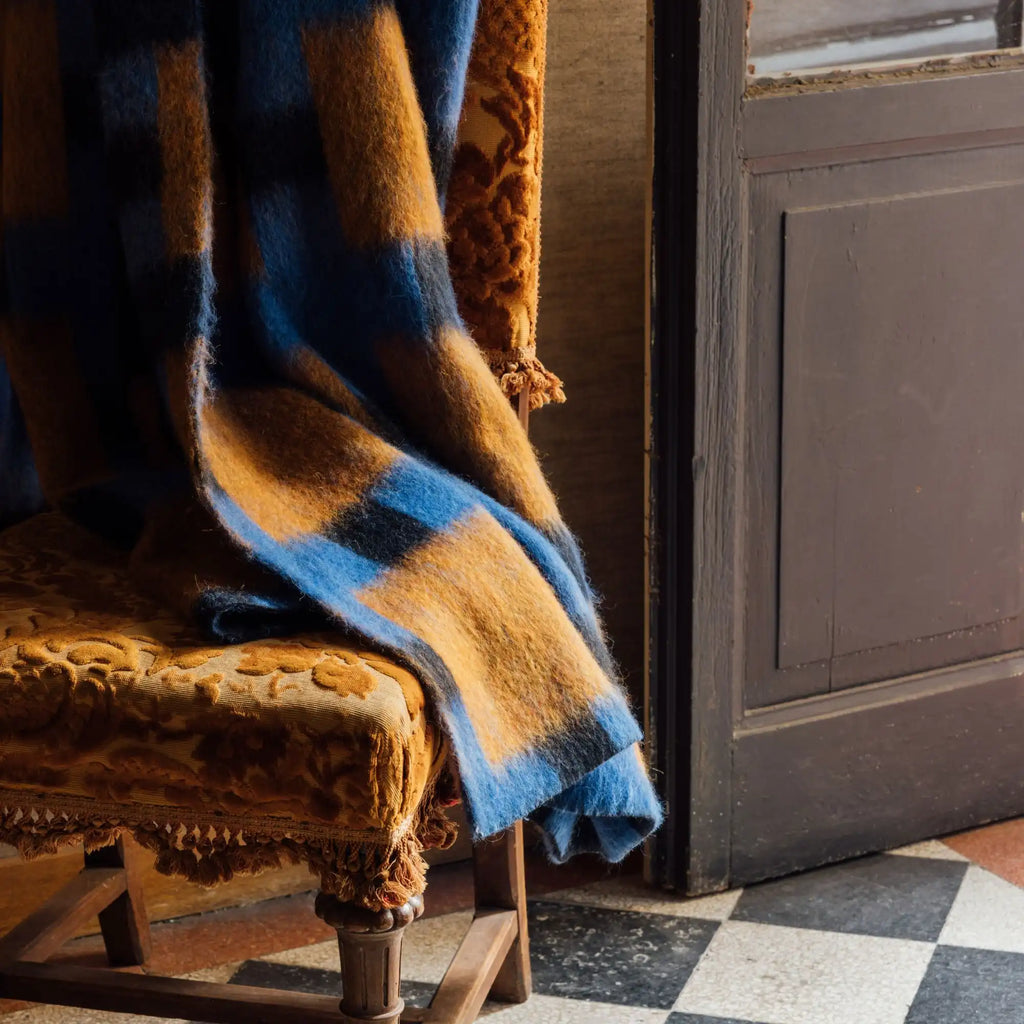 Layered Fall Foliage Oversized Blanket, a cozy blue and orange plaid wool throw draped over a vintage ornate chair. Perfect for adding warmth and rustic charm to any living space or bedroom decor.