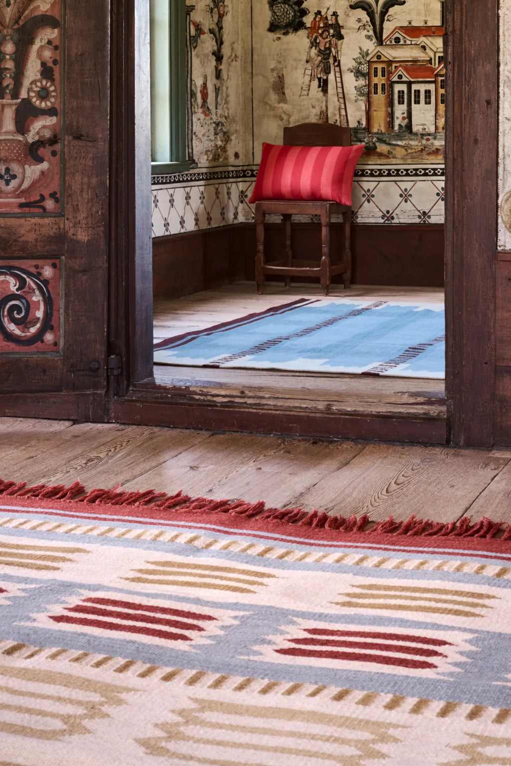 Layered Hilma Wool Rug, a handwoven natural wool rug with a light blue, cream, and red ikat pattern and fringed edges. Perfect for adding global-inspired warmth and texture to any living space.