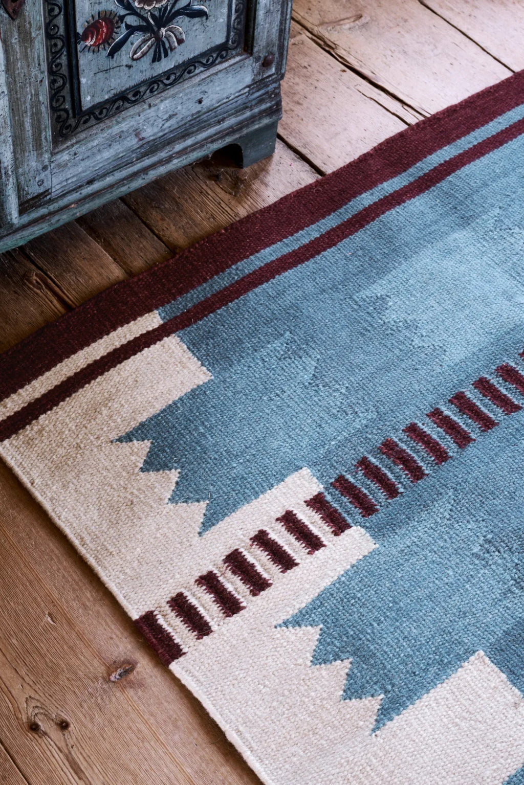 Layered Iris Wool Rug, a hand-woven blue and off-white patterned area rug with burgundy striped accents on a rustic wooden floor. Perfect for adding a cozy, bohemian touch to any living space or be...