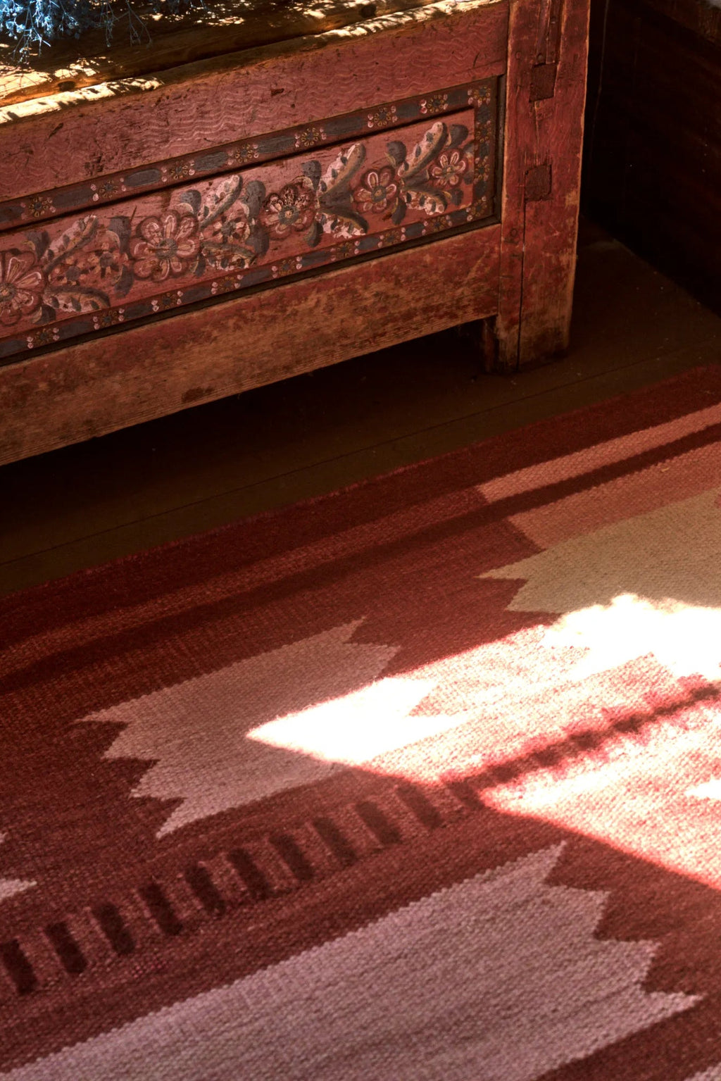 Layered Iris Wool Rug - Raspberry, a handwoven wool rug featuring a striking Southwestern-inspired geometric pattern in shades of raspberry red and light pink, partially illuminated by sunlight, ne...