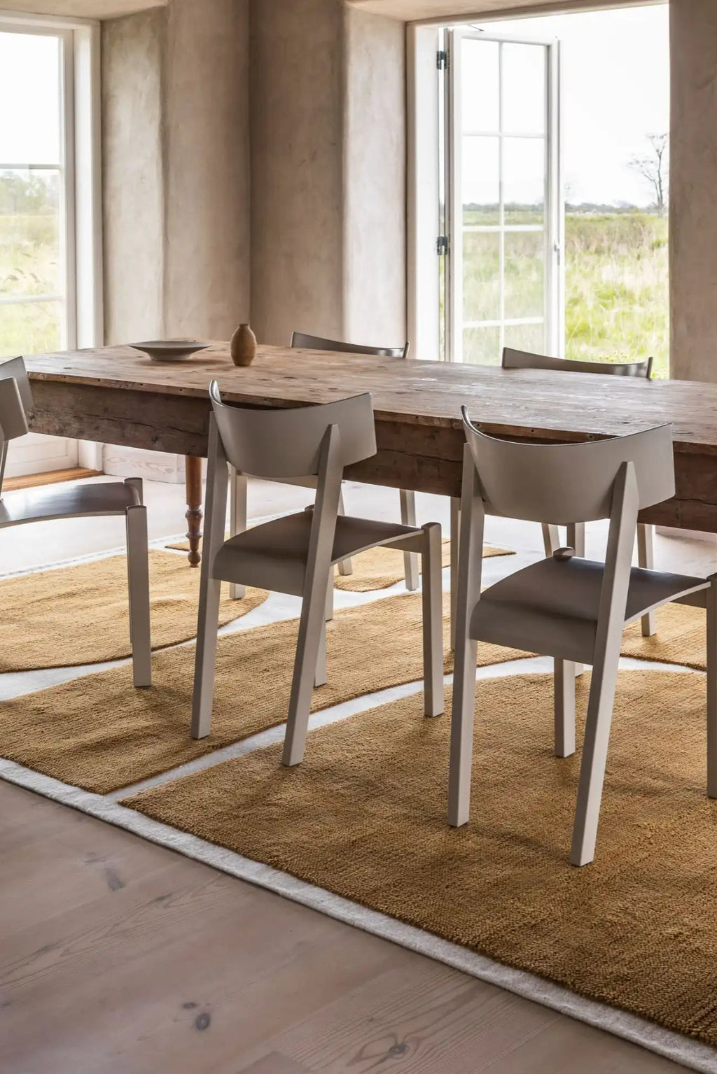 Layered Gotland Rauk Ullmatta Wool Rug, a textured natural tan wool runner rug with a subtle cream border, providing warmth under a rustic wooden dining table and modern chairs. Perfect for adding...