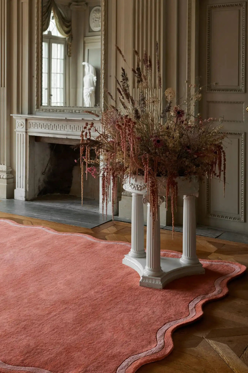 Layered Scallop Wool Rug, a luxurious coral pink area rug with an elegant, wavy scalloped border in lighter pink, on a wooden parquet floor. Perfect for adding a touch of sophisticated warmth to li...