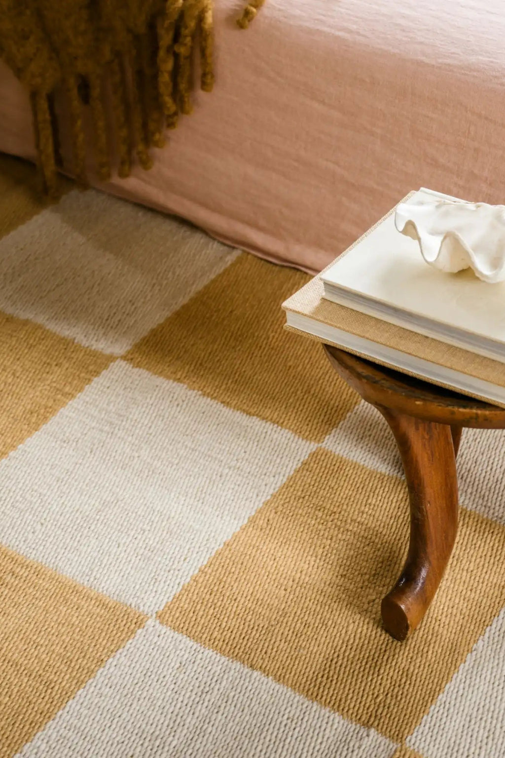 Layered Chess Wool Rug, an ivory and goldenrod yellow checkered wool area rug with a woven texture, partially visible under a bed with a small wooden side table. Perfect for adding a warm, geometri...