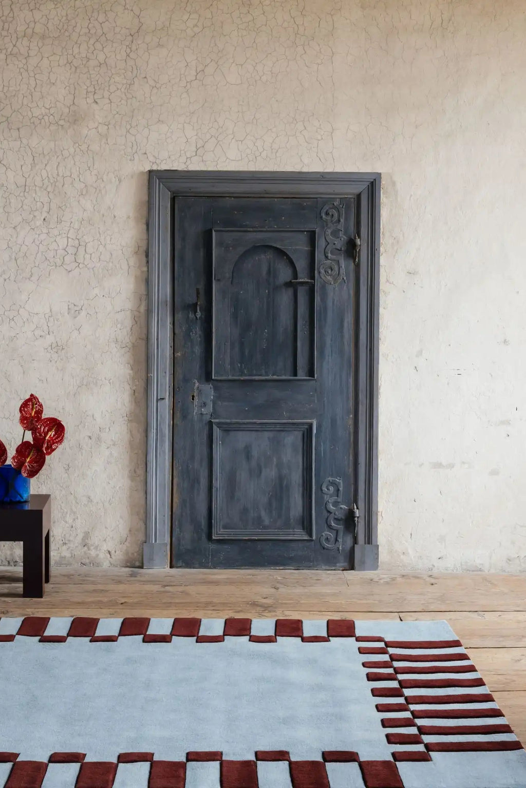Layered Frame Wool Rug, a modern light blue wool rug with a striking chunky red checkerboard border design, on a rustic wooden floor. Perfect for adding a contemporary accent to a living room or be...