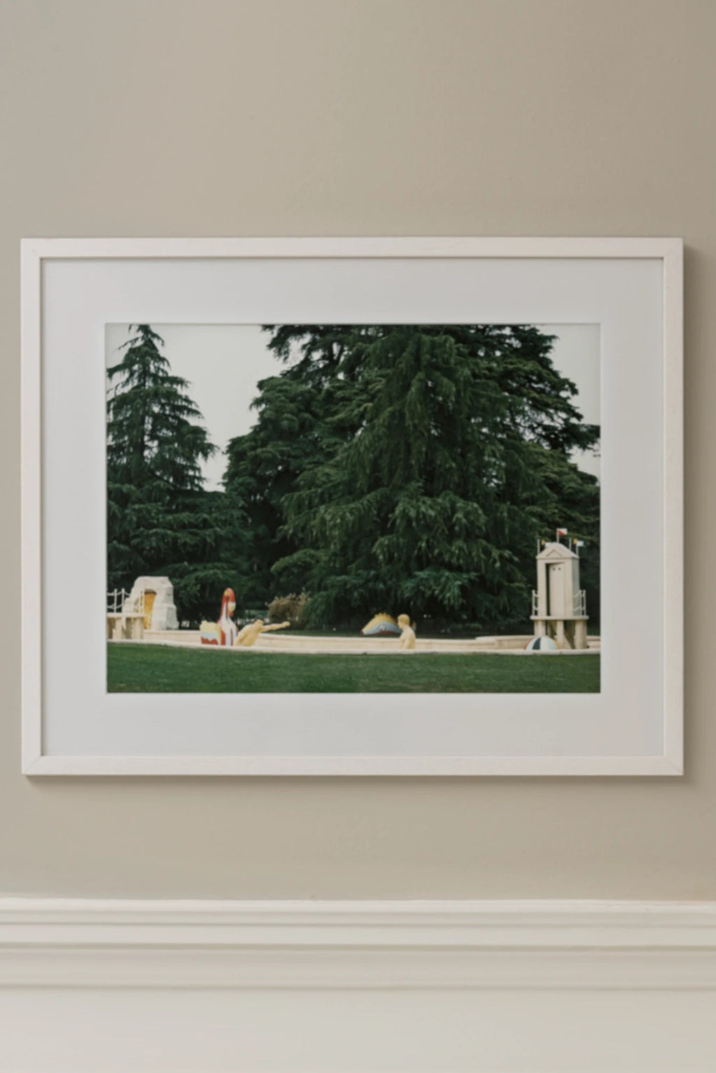 The Poster Club Biennale, a fine art photography print capturing a serene outdoor park scene with towering deep green evergreen trees and a vibrant green lawn. The image features cream-colored playground structures and small figures, presented with a crisp white mat and a clean white minimalist frame. This modern wall art piece is perfect for contemporary living rooms, minimalist bedrooms, or a unique addition to a gallery wall, offering a tranquil yet whimsical aesthetic.