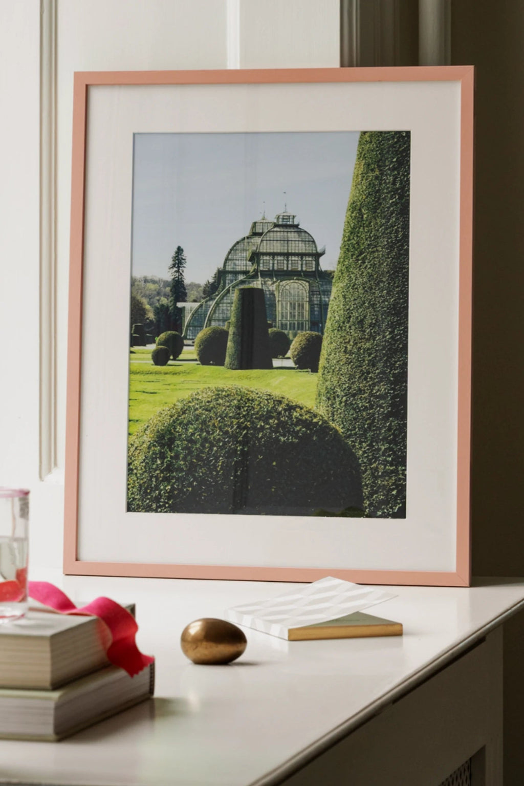 The Poster Club Topiary, a contemporary architectural garden photography print featuring a grand greenhouse surrounded by meticulously sculpted green topiary trees and lush grass, framed in a delicate pink wood frame with a crisp white mat. This elegant wall art is perfect for botanical-inspired gallery walls or as a sophisticated accent in modern living rooms and bedrooms.