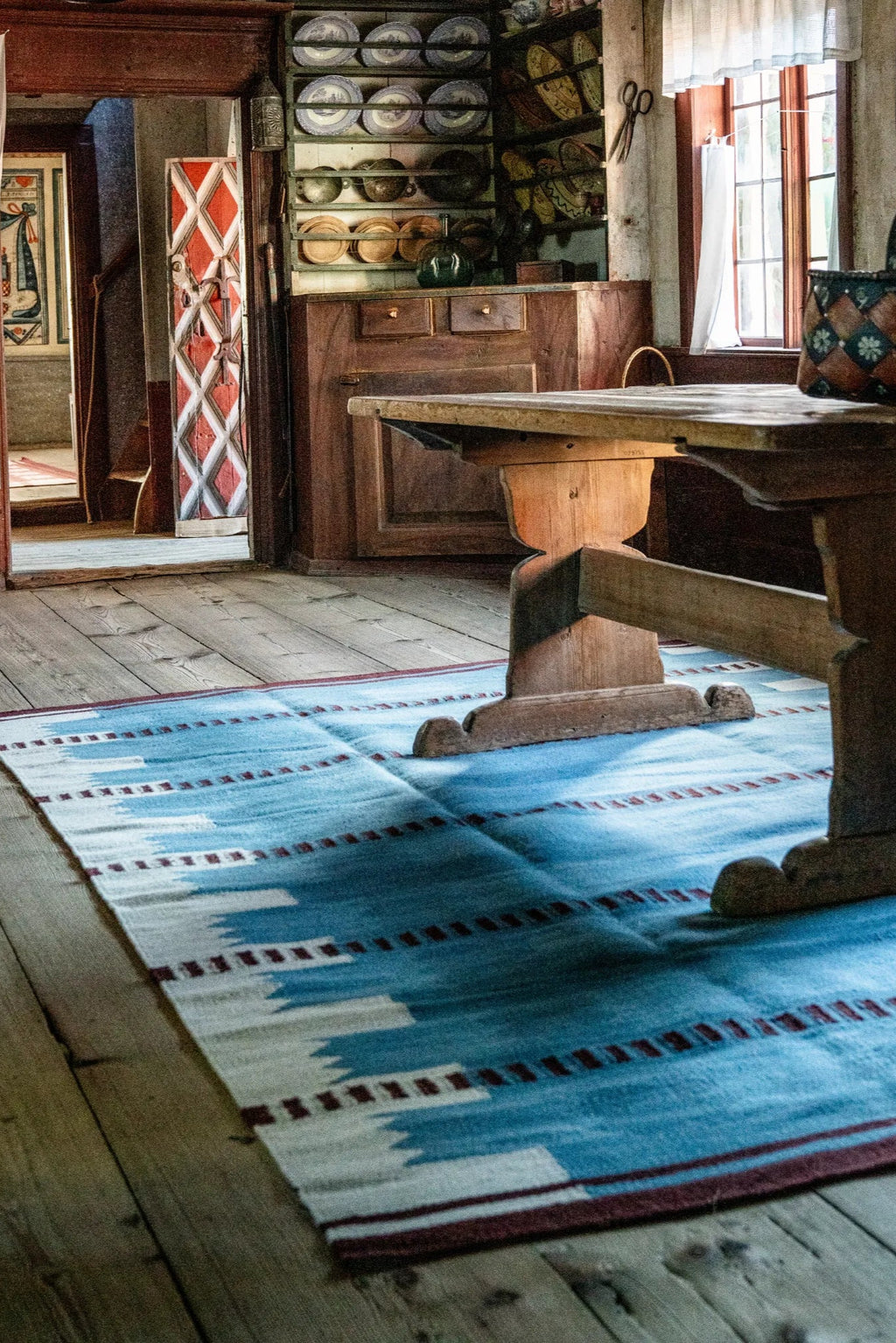 Layered Iris Wool Rug, a hand-woven blue and white patterned flatweave area rug with burgundy accents, laid on a rustic wooden floor. Perfect for adding a touch of Scandinavian folk art style to an...