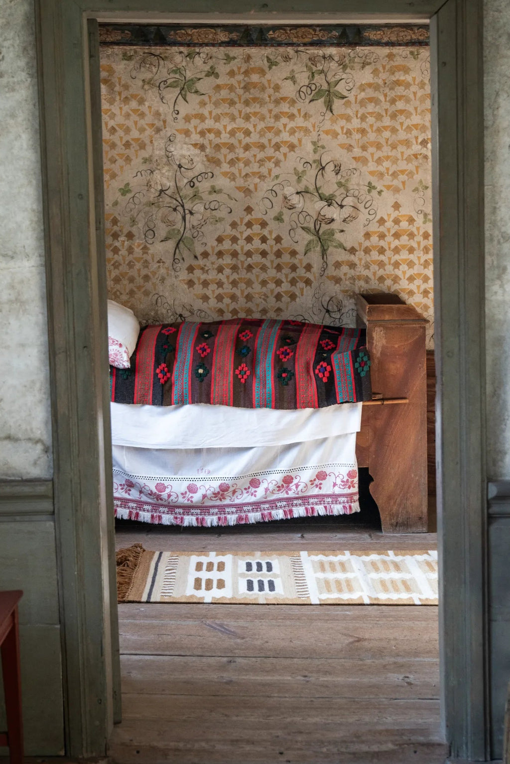 Layered Hilma Wool Rug, a handwoven brown, beige, and white patterned wool rug with a geometric house design and fringed edges, visible on a rustic wooden floor. Perfect for adding a cozy, natural...