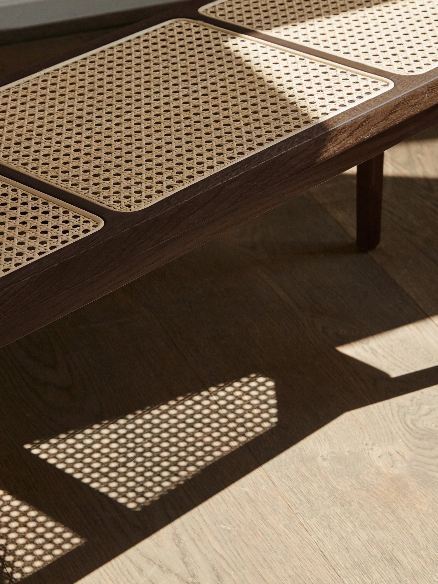 NORR11 Le Roi Bench, a premium dark brown oak wood frame with natural light beige cane webbing seats, shown in dappled sunlight on wood flooring. Ideal for sophisticated living spaces.