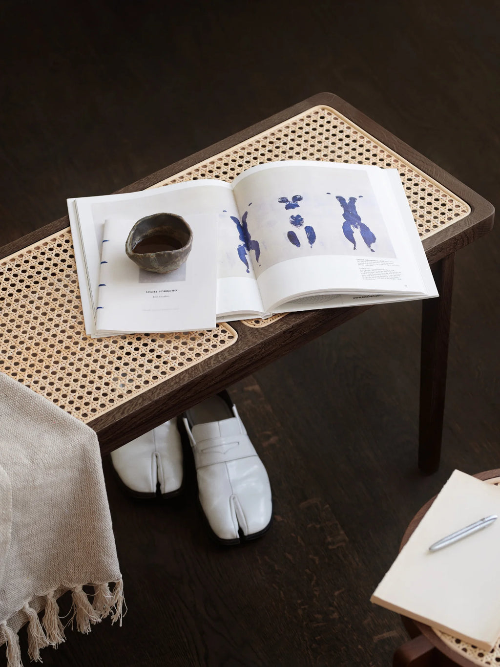 NORR11 Le Roi Bench, a luxurious dark stained oak wood bench with natural cane webbing top, adorned with an open book and ceramic cup, enhancing any modern interior.