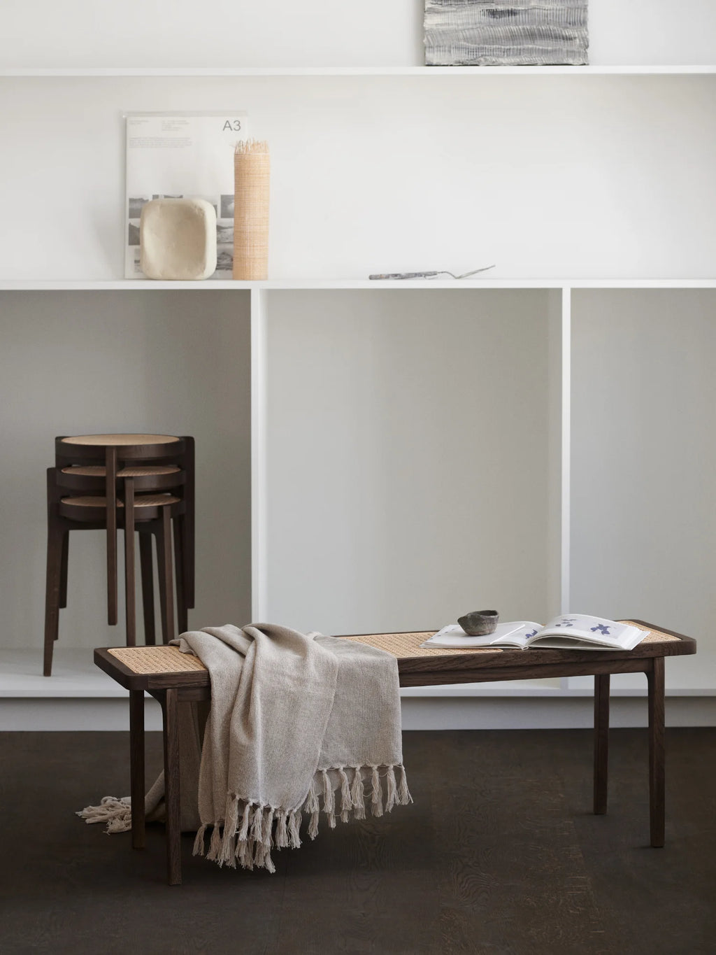 NORR11 Le Roi Bench. Lifestyle shot of a dark brown wooden bench with a natural cane woven seat, styled with a light beige fringed throw. A luxury furniture piece for any modern interior.