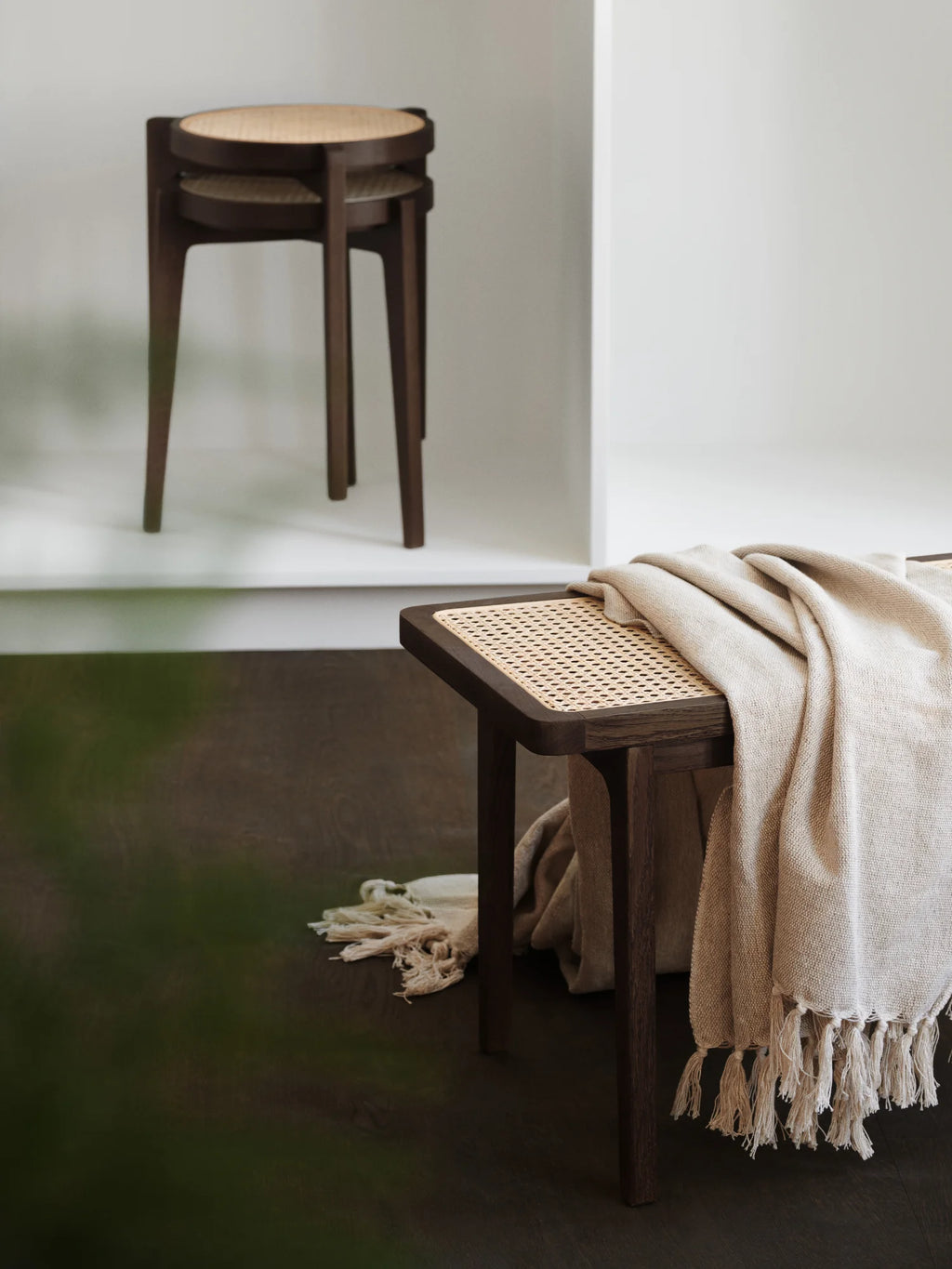 NORR11 Le Roi Bench, a dark brown solid oak and natural cane webbing luxury bench, styled with a light beige knit throw blanket with fringe, sits on a dark wood floor in a modern room. Perfect for elegant home decor.