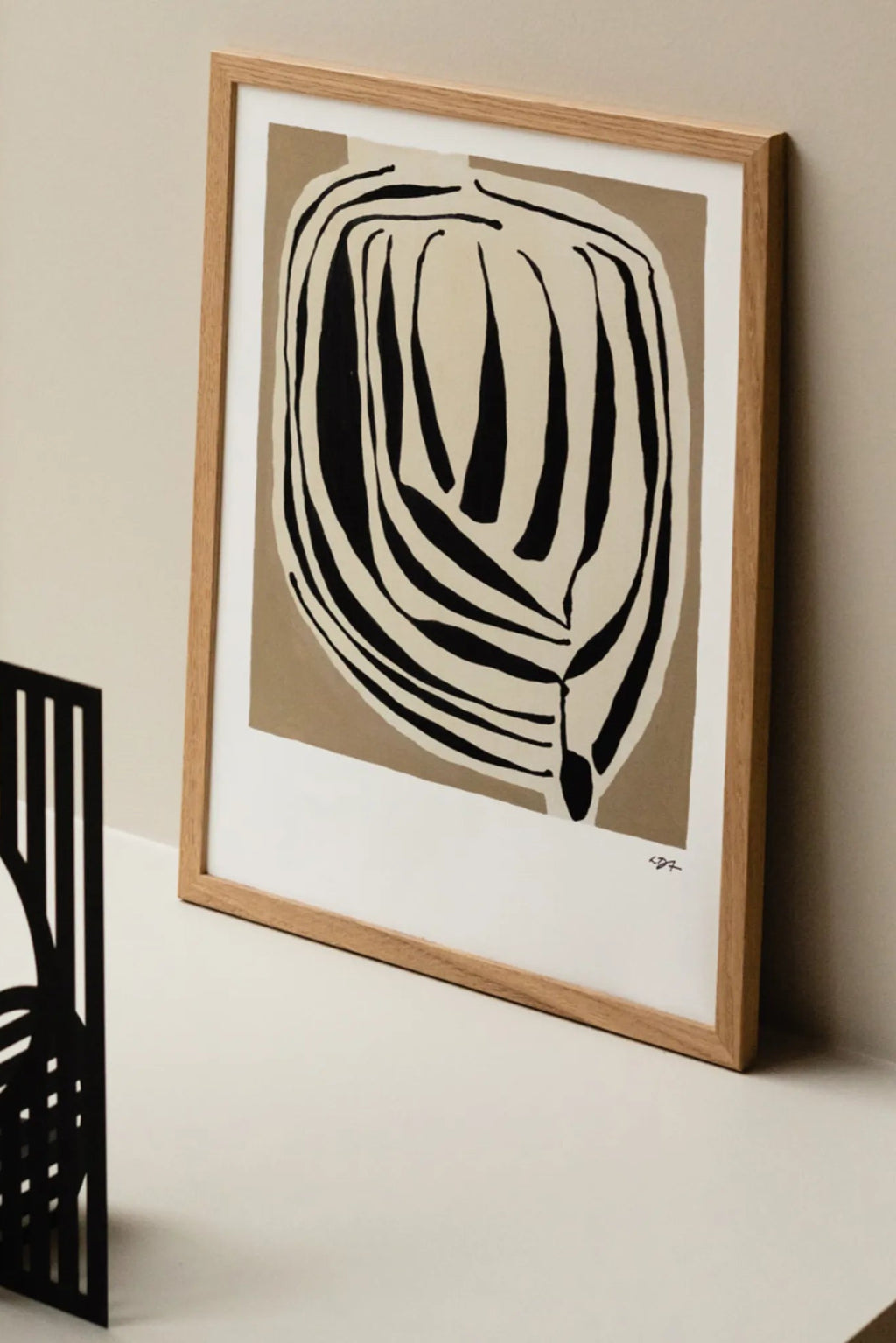 The Poster Club Layered, an abstract art print featuring bold black organic shapes layered over a warm beige background, contrasted by an off-white border. It is elegantly presented in a light natural oak frame with a crisp white mat. Perfect as minimalist wall art for a contemporary gallery wall or organic modern living room decor.