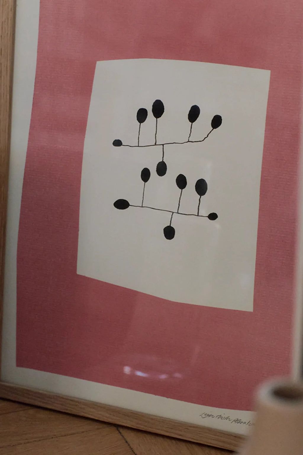 The Poster Club Pink Surrender, a minimalist abstract art print featuring delicate black organic shapes on an off-white center, surrounded by a dusty rose pink background with a subtle paper texture. This contemporary wall art is presented in a natural oak frame with a crisp white mat. Ideal for Scandinavian living rooms, organic modern bedrooms, or a stylish gallery wall accent.