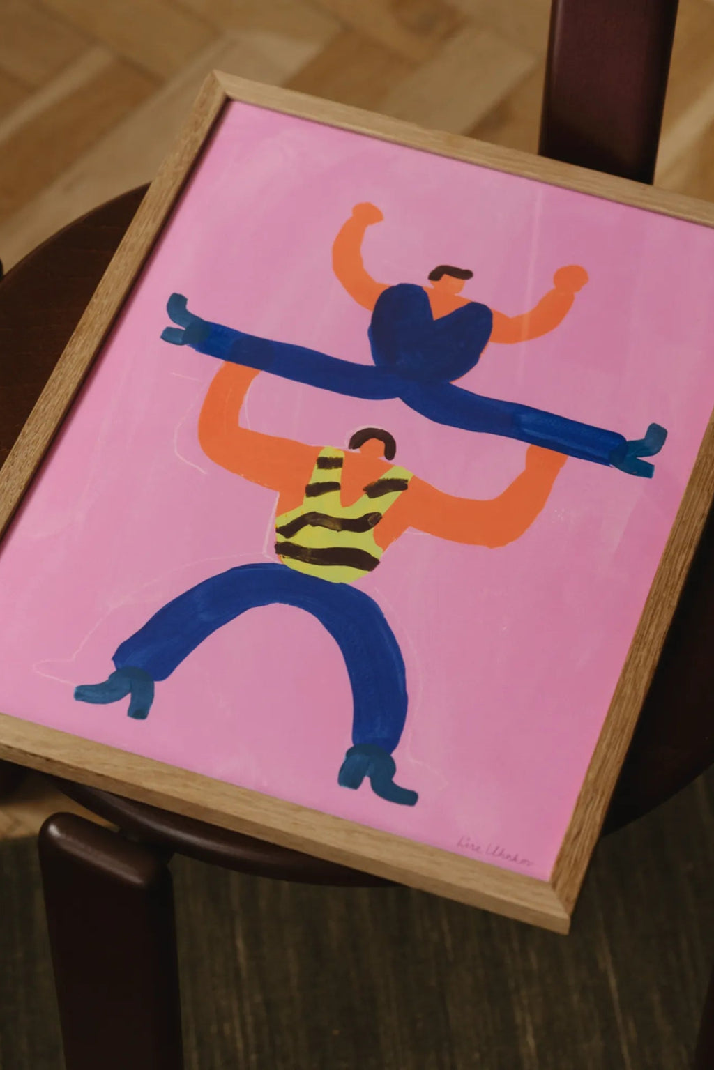 The Poster Club I will super carry You, a vibrant contemporary art print featuring an abstract figurative design with two figures in royal blue and bright orange on a bold pink background, one holding the other overhead. This playful graphic poster is presented in a light natural oak frame, ideal as colorful wall art for modern living rooms, kids' bedrooms, or a lively gallery wall.