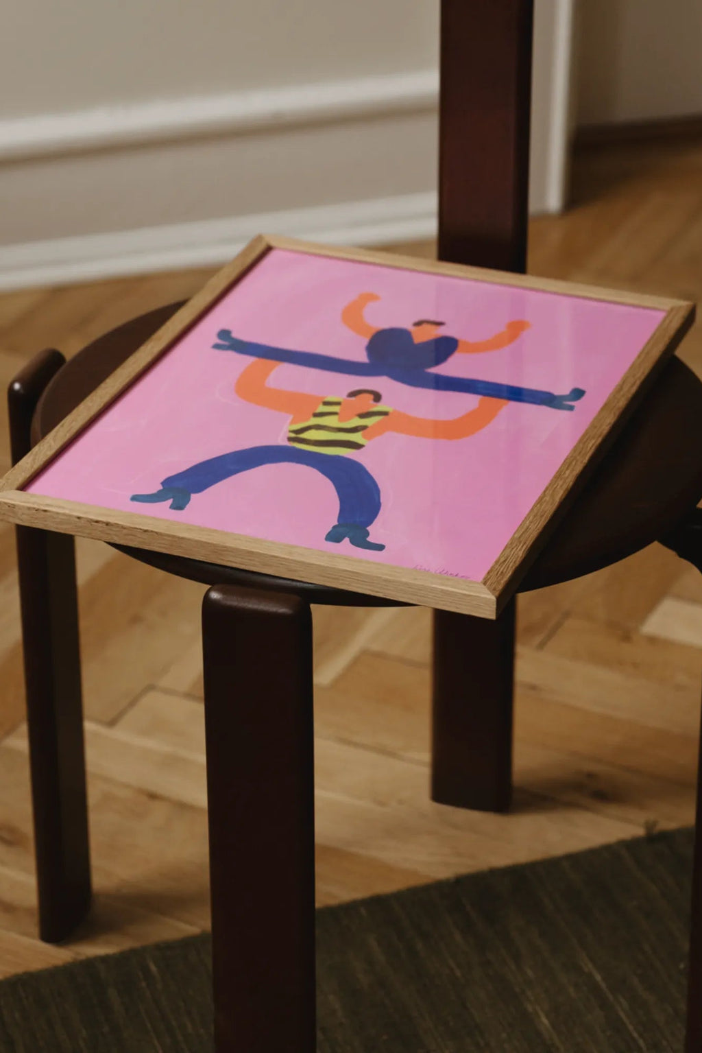 The Poster Club I will super carry You, a vibrant abstract art print featuring two playful, stylized figures in dark blue and orange, one wearing a yellow striped shirt, set against a bold pink background. This colorful contemporary poster, framed in light natural oak, offers a cheerful folk art style. Ideal as modern wall art for a gallery wall, a fun bedroom accent, or vibrant nursery decor.