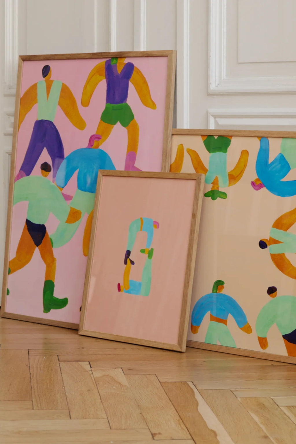 The Poster Club Position 0, a vibrant abstract art print featuring graphic, colorful human-like figures in shades of yellow, purple, green, and blue against a soft pink background, enclosed in a natural oak frame. This modern framed poster is perfect as playful wall art for contemporary living rooms, bedrooms, or creative gallery walls.