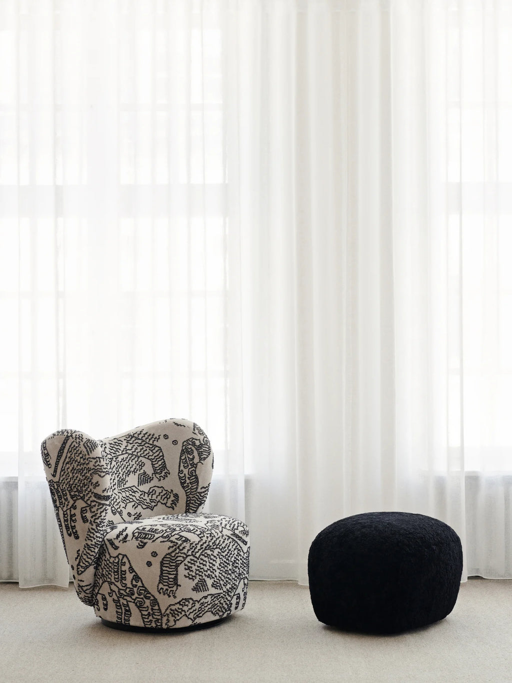 NORR11 Little Big Pouf, a small, round, textured black pouf sits on a light carpet next to a patterned armchair. This elegant, modern accent piece adds comfort and a stylish touch to any living space for discerning homeowners.
