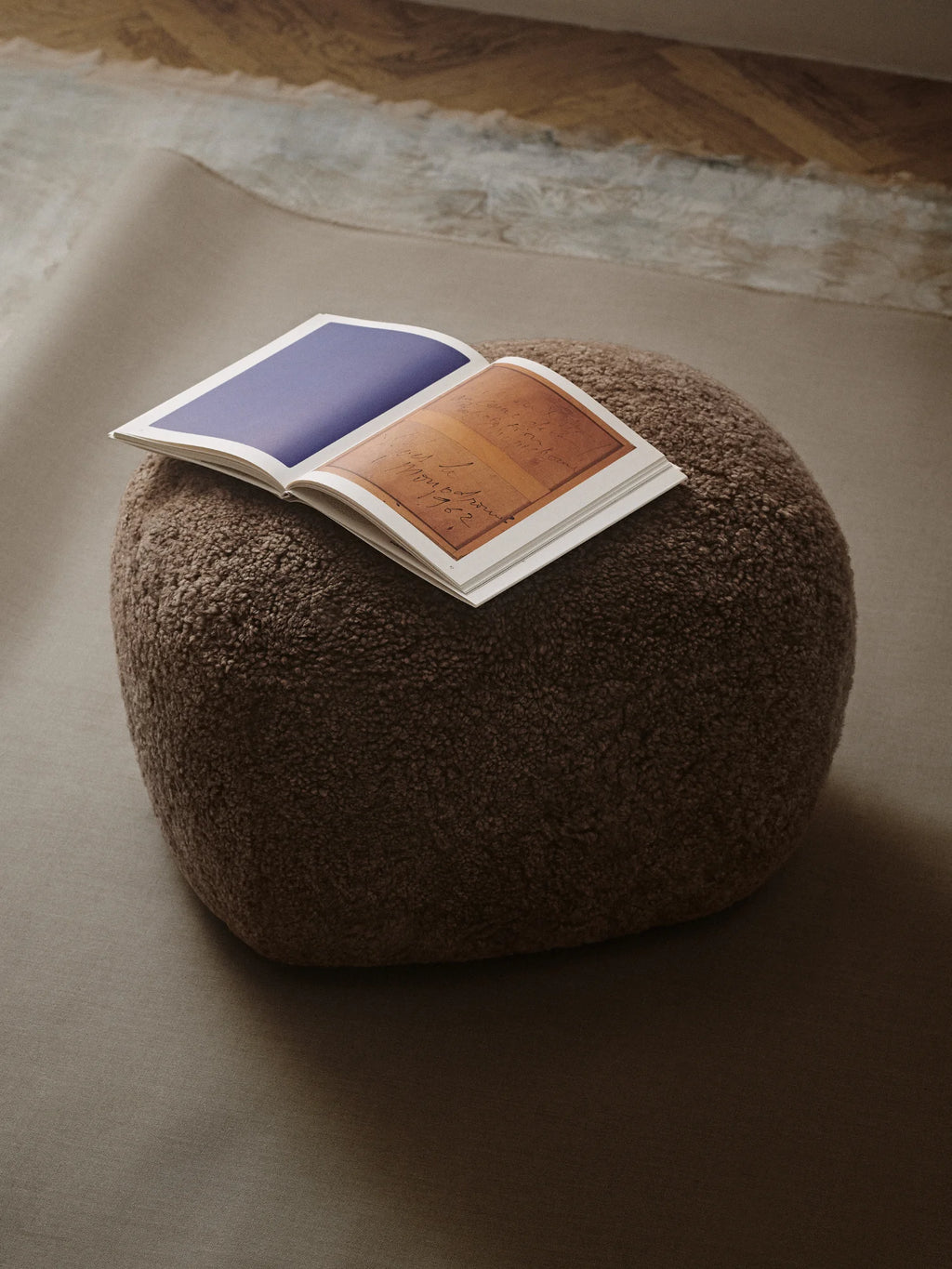 NORR11 Little Big Pouf, a premium and stylish pouf in a deep brown textured boucle material. An open book with artistic pages rests on its plush surface, adding a touch of modern elegance. Perfect as versatile seating or an elegant footrest for sophisticated living spaces.