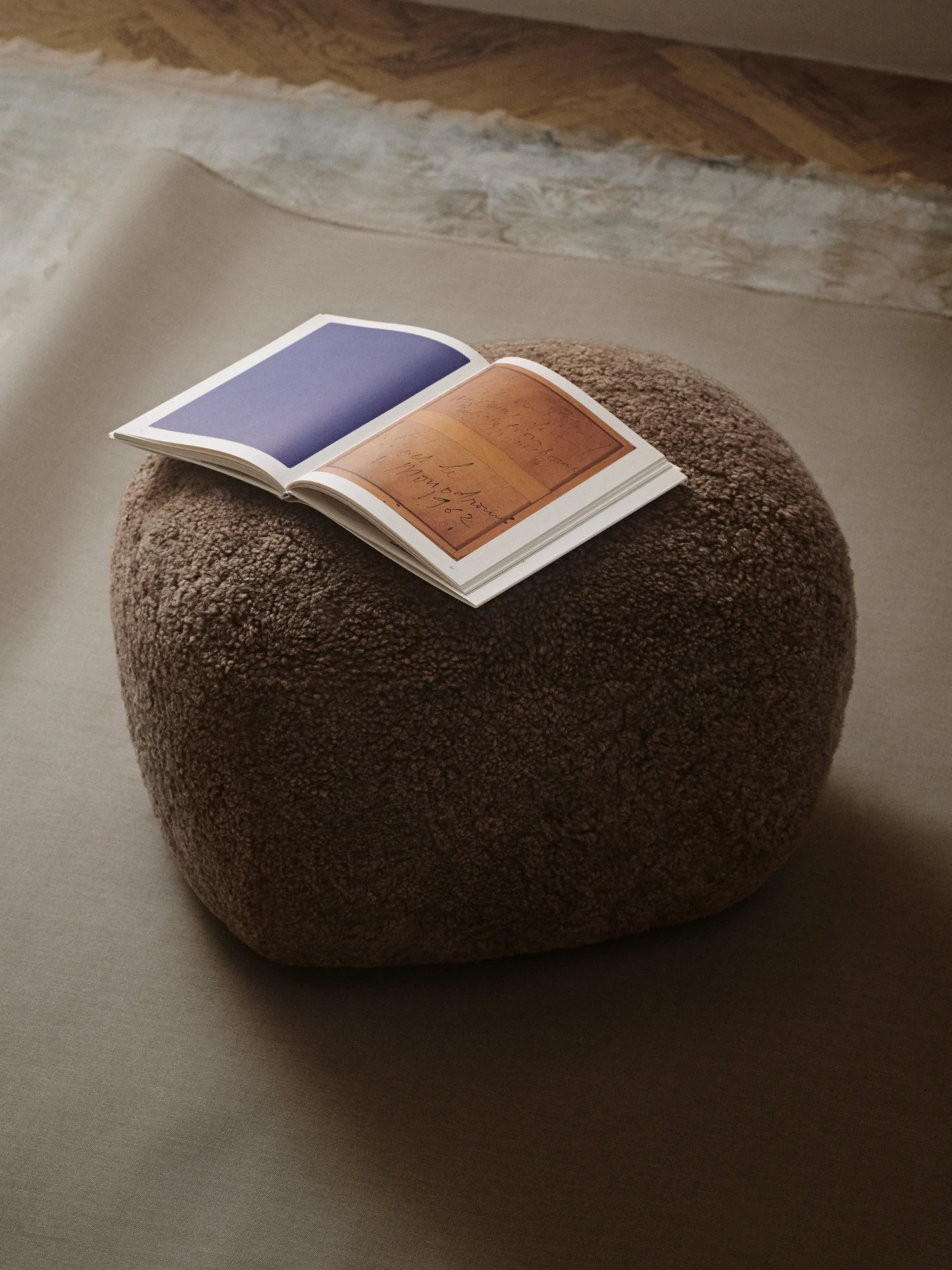 NORR11 Little Big Pouf, a premium and stylish pouf in a deep brown textured boucle material. An open book with artistic pages rests on its plush surface, adding a touch of modern elegance. Perfect as versatile seating or an elegant footrest for sophisticated living spaces.