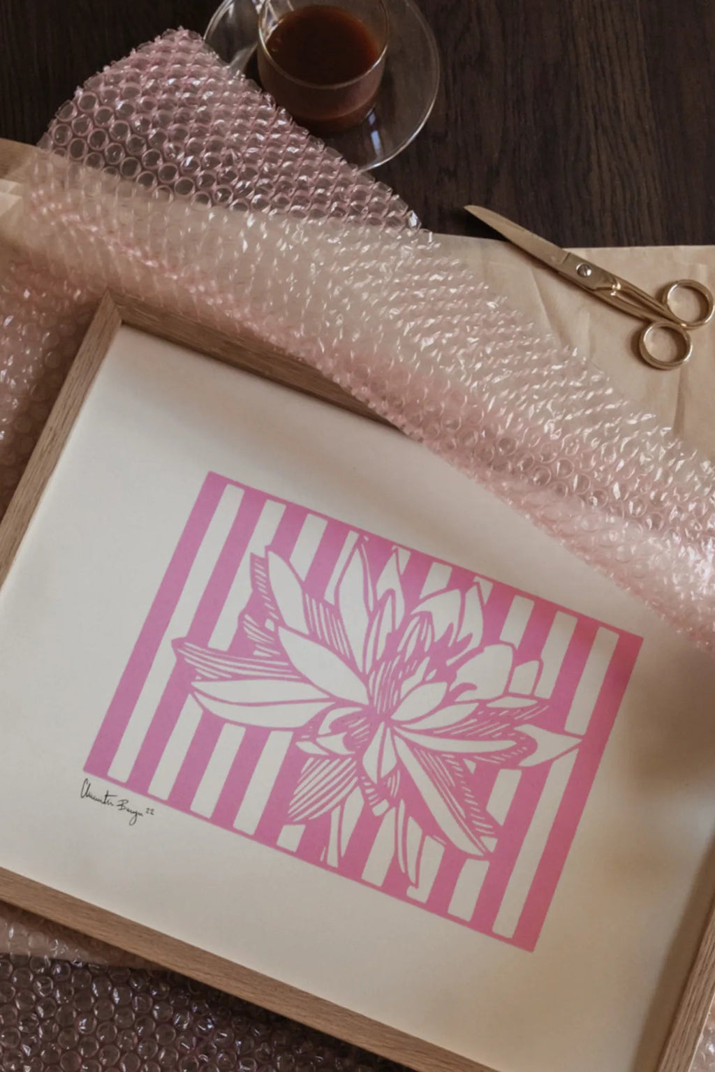 The Poster Club Lotus No 01, a modern graphic art print featuring a stylized pink lotus flower against a crisp white background with bold vertical pink stripes, presented in a light natural oak frame. This contemporary pink floral poster is perfect for a minimalist bedroom accent or a vibrant gallery wall in modern living rooms.
