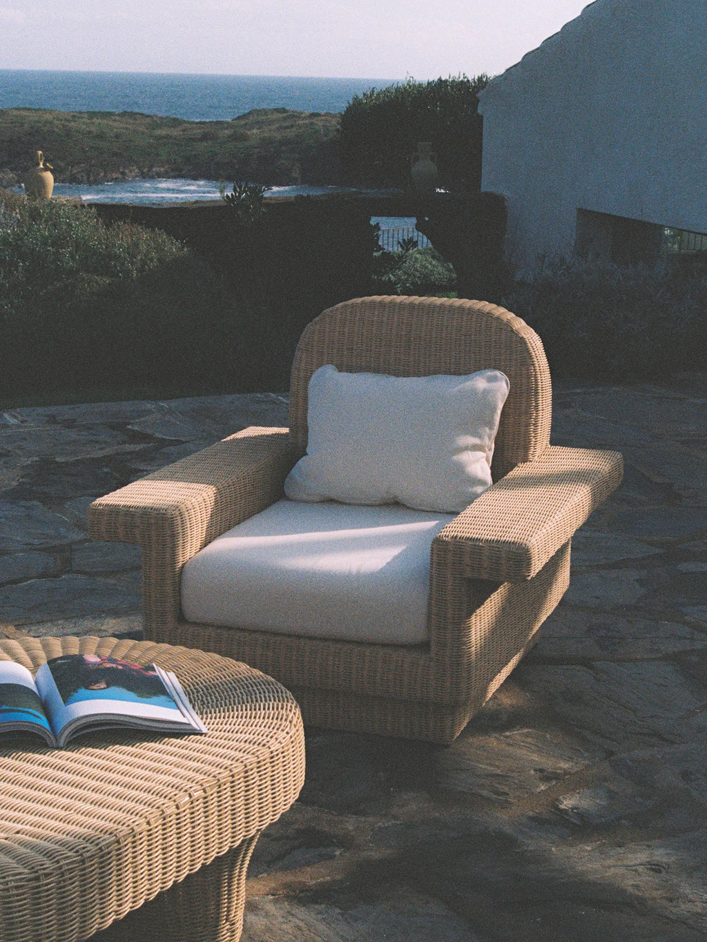 Heaps & Woods Lola Lounge Chair w/ Armrest, a natural woven rattan lounge chair with a gently curved backrest, wide flat armrests, and plush off-white cushions. This outdoor armchair features a rustic yet elegant silhouette, crafted from light brown wicker with a cozy, inviting texture. Perfect for coastal patios, Mediterranean terraces, or organic modern outdoor living spaces.