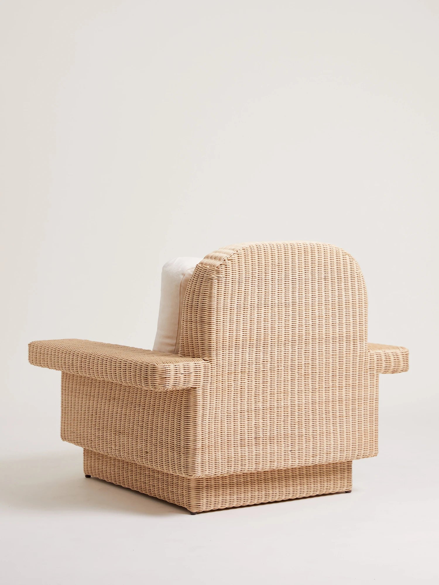 Heaps & Woods Lola Lounge Chair w/ Armrest, a natural woven rattan lounge chair featuring a distinctive rounded backrest, wide flat armrests, and a sturdy, block-style plinth base. Crafted from light beige rattan with a textured weave, this contemporary armchair includes a subtle white cushion. Ideal as an organic modern accent chair for living rooms, sunrooms, or coastal-inspired spaces.