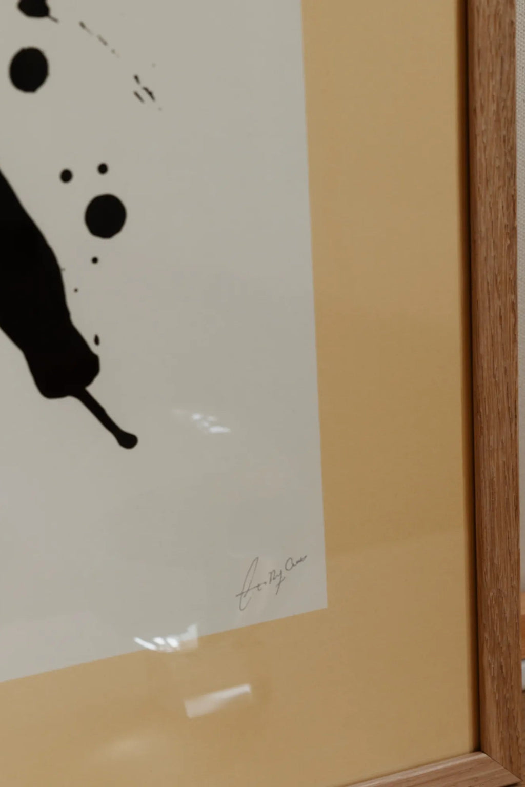 The Poster Club Imagine, a striking abstract art print featuring dynamic black ink splatters and organic marks against a clean white background, expertly framed with a warm pale yellow mat and a light natural oak frame.