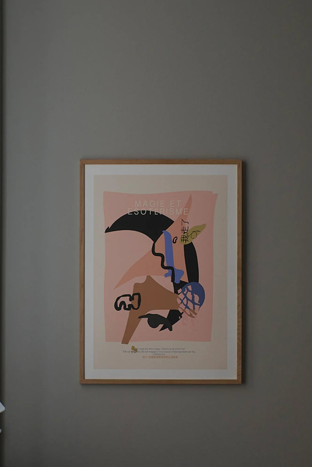 The Poster Club Magie, a modern abstract art print featuring bold graphic shapes in black, royal blue, warm brown, and olive green against a soft blush pink background, with the French text "MAGIE ET ESOTERISME." This contemporary poster is framed in natural oak with a crisp white mat, making it ideal wall art for a minimalist living room, Scandinavian bedroom, or modern gallery wall.