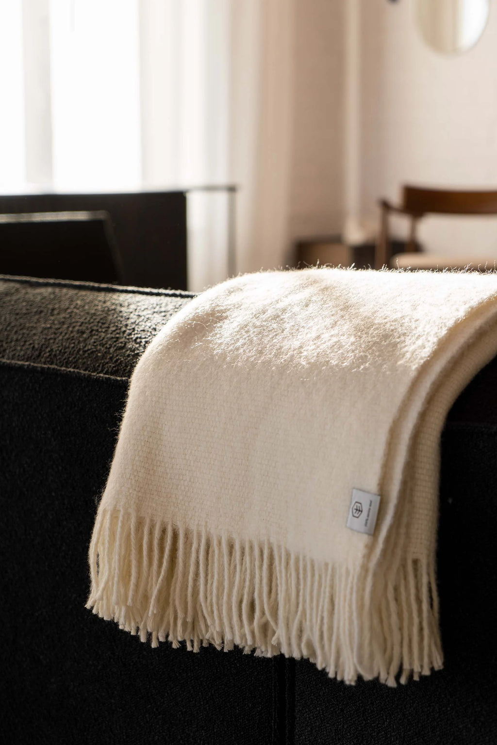 TAMO Meri Blanket Throw: A luxurious, creamy white woven throw with visible fringe and brand tag, draped over a dark sofa in a sunlit room. Perfect for cozy home decor.