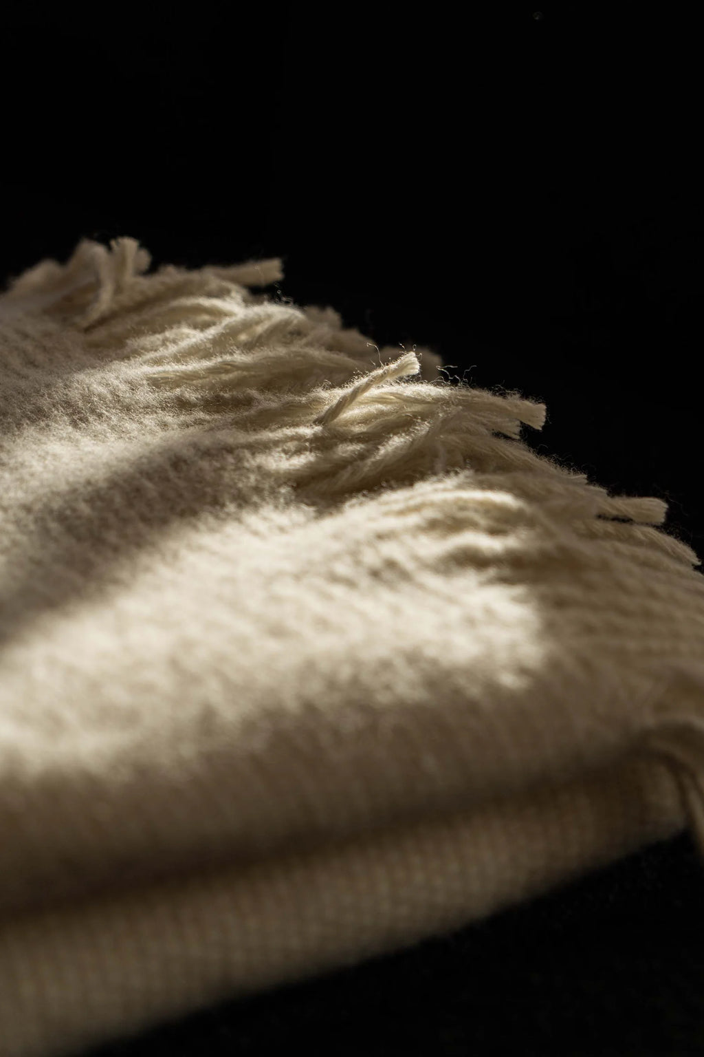 TAMO Meri Blanket Throw, a close-up of a premium cream-colored woven fabric with fringed edges, highlighted by a diagonal beam of light on a black background. Ideal for cozy home decor.
