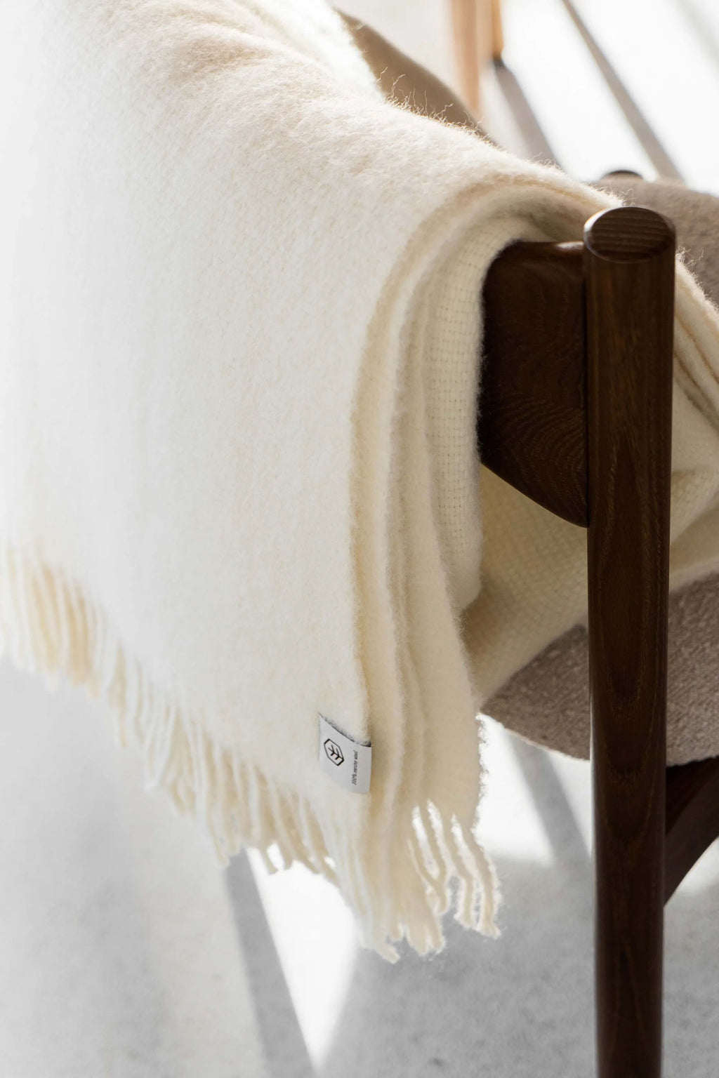 TAMO Meri Blanket Throw: Fluffy, creamy white 100% merino wool blanket with fringed edges draped over a dark wood chair, highlighting its luxurious texture. Ideal for cozy home decor.