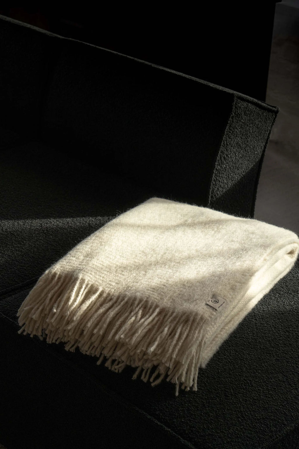 TAMO Meri Blanket Throw, a premium cream-colored wool throw with visible fringe details, folded neatly on a dark gray couch, highlighted by natural sunlight for a cozy feel. Perfect for a luxurious home accent.