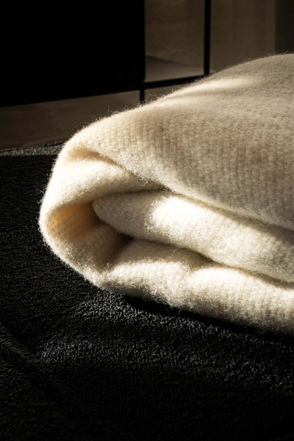 TAMO Meri Blanket Throw: Close-up of a luxuriously soft, ivory white, folded knitted throw with visible fuzzy texture, resting on a dark, textured surface, illuminated by natural light. Ideal for cozy home decor.