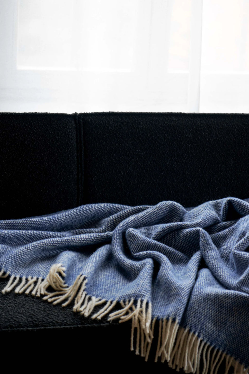 TAMO Meri Blanket Throw, a premium blue and white flecked woven wool throw with cream fringe, draped on a dark sofa. Ideal for cozy living room decor.