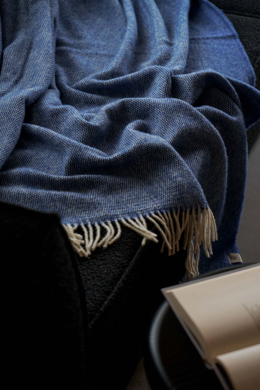 TAMO Meri Blanket Throw, a premium blue woven fabric with subtle white flecks, draped over a dark chair with off-white fringed edges. Ideal for cozy home decor.