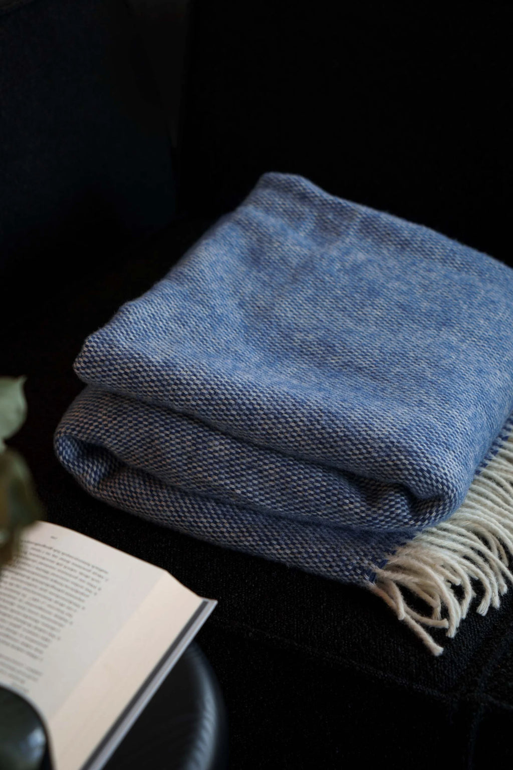 TAMO Meri Blanket Throw, a soft, premium chambray blue woven blanket with visible white fringes, folded neatly on a dark sofa next to an open book. Ideal for cozy living spaces.