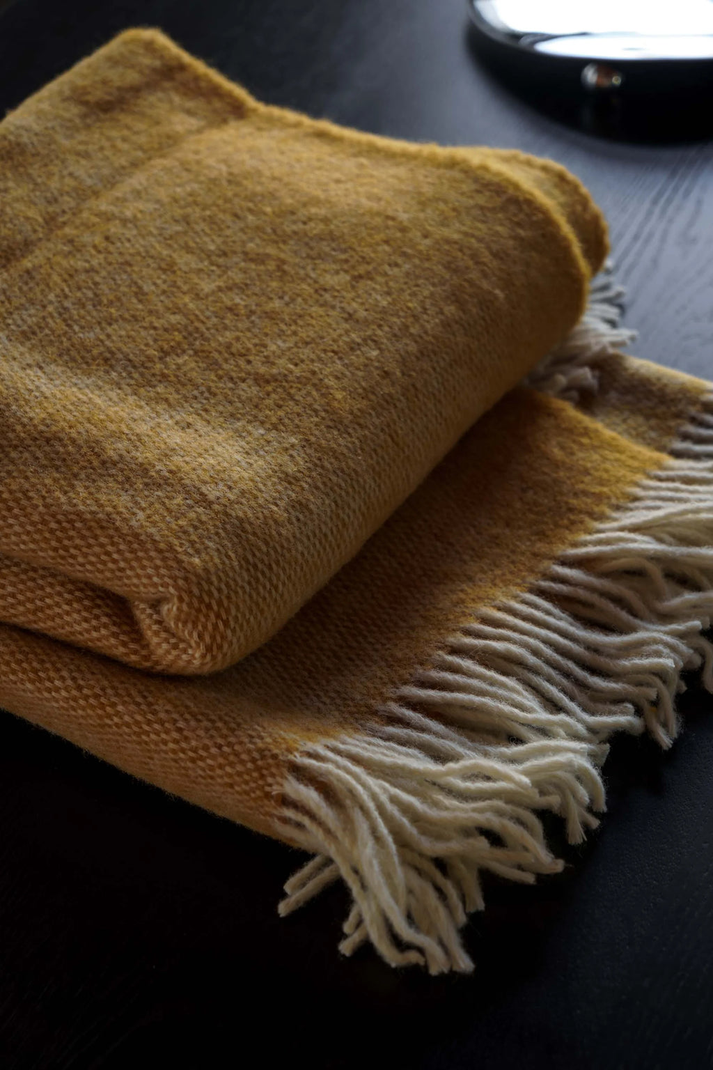 TAMO Meri Blanket Throw, a folded golden-yellow premium woven knit blanket with cream-colored fringe, displayed on a dark surface. Perfect for adding cozy warmth to any room.