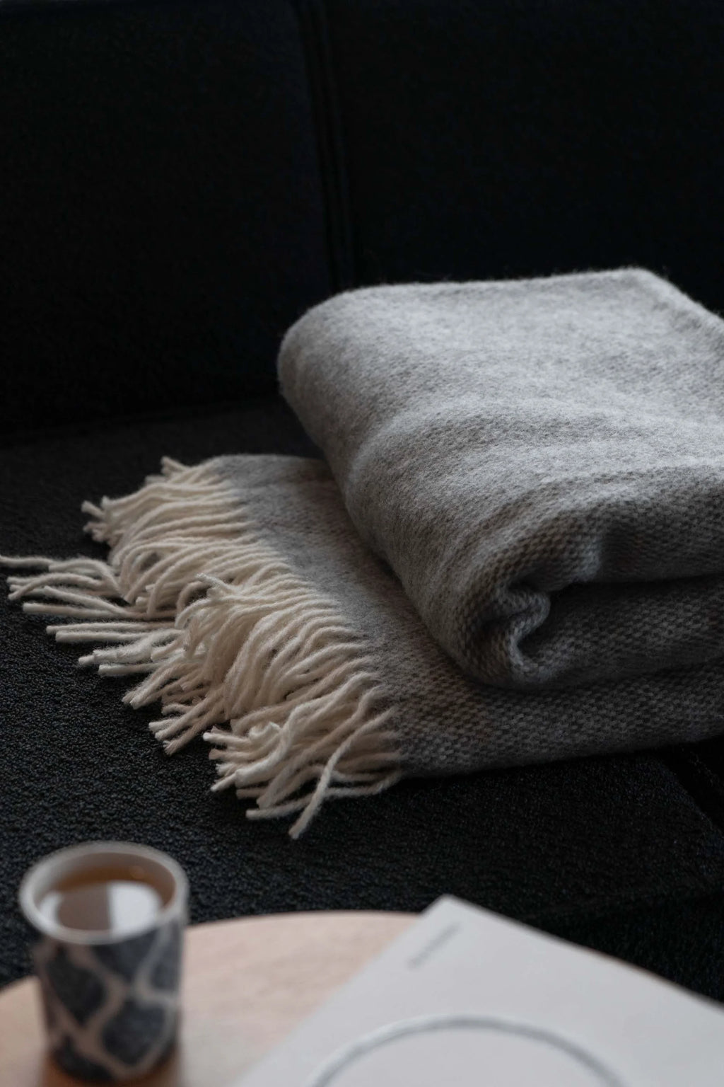 TAMO Meri Blanket Throw: Folded luxurious light grey woven knit throw with cream fringe detailing, styled on a black sofa. Ideal for cozy home decor.