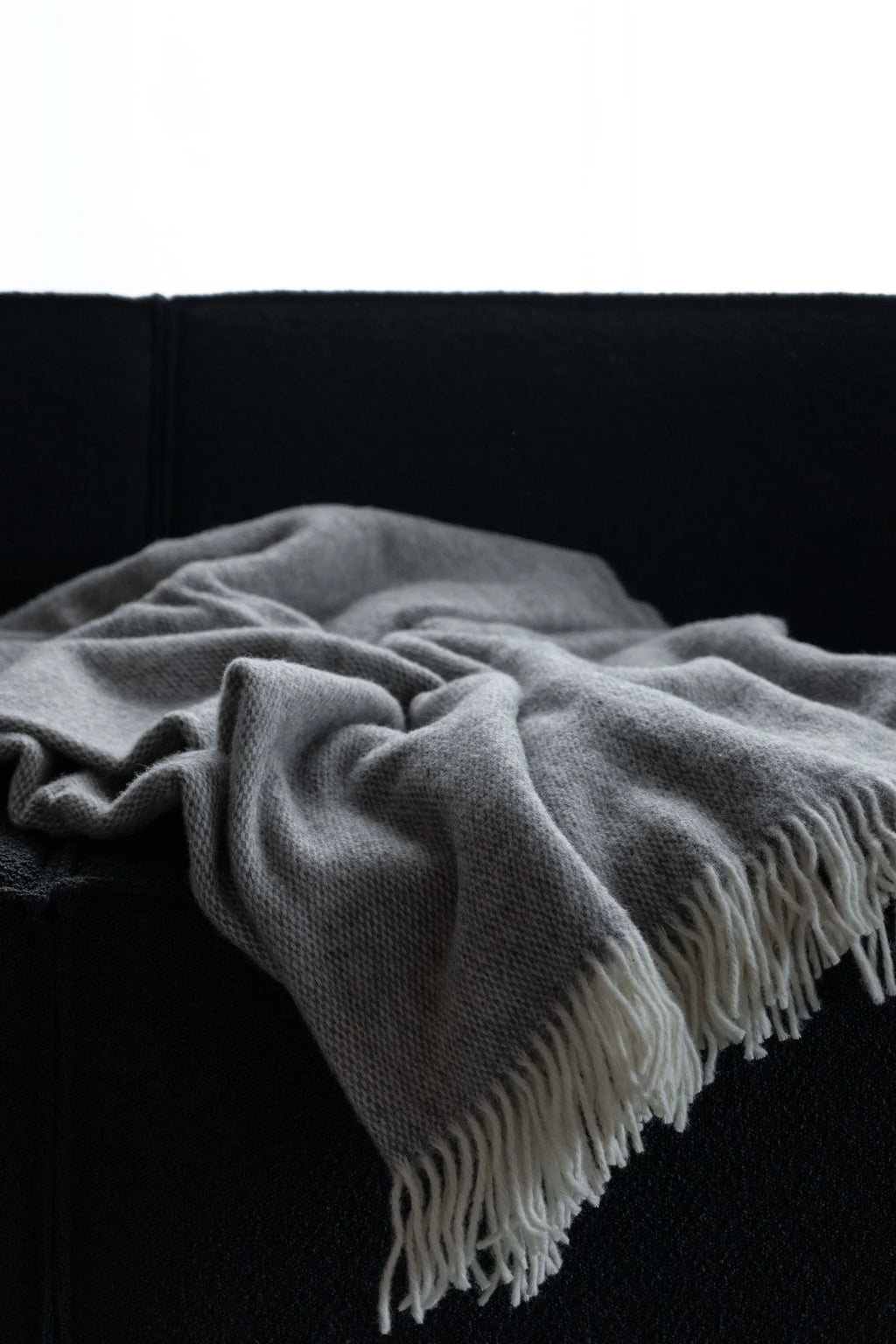 TAMO Meri Blanket Throw: A soft, luxurious light gray knitted blanket detailed with white fringe, casually draped over a dark sofa. Perfect for adding cozy comfort to any living space.