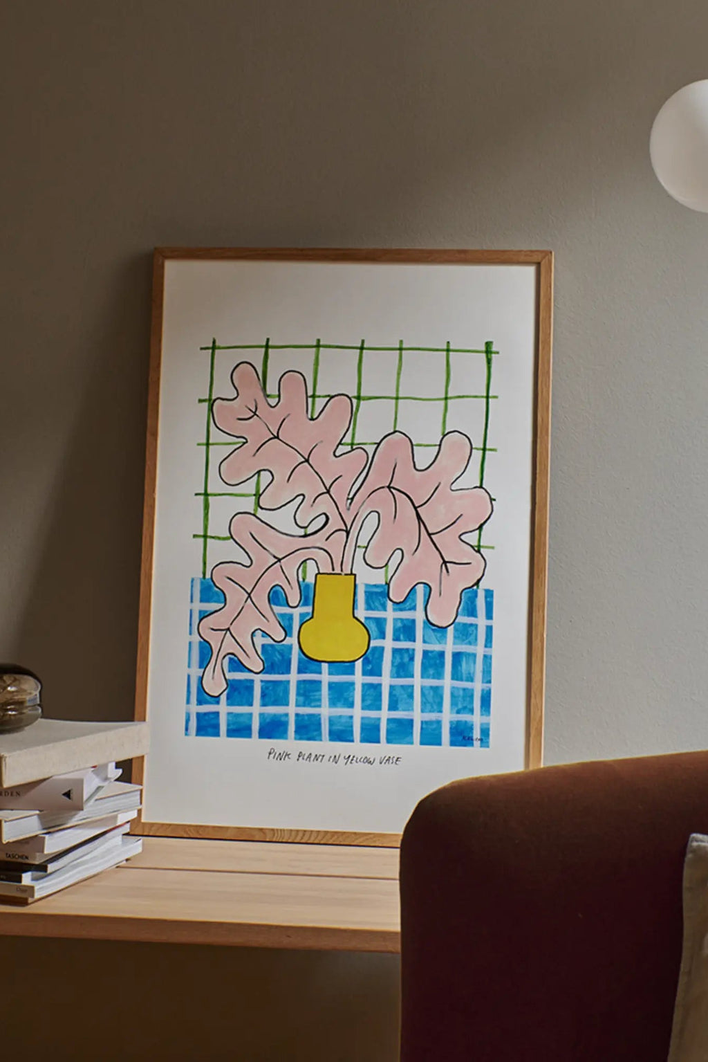 The Poster Club Pink Plant in Yellow Vase, a contemporary graphic art print featuring bold pink organic plant leaves in a bright yellow vase, set against a vibrant blue checkered pattern and a subtle green grid background. This colorful minimalist poster is framed in natural oak wood, ideal as playful wall art for modern living rooms, bedrooms, or creative home office spaces, adding a pop of color and abstract botanical style.
