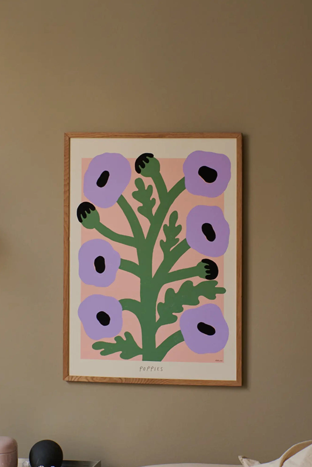 The Poster Club Purple Poppies, a modern abstract botanical art print featuring simplified lilac purple poppy flowers with black centers and deep green stems and leaves, set against a soft pale pink background. The paper print is housed in a natural light oak wood frame with a crisp white mat. Ideal as contemporary wall art for a minimalist living room, Scandinavian bedroom, or playful nursery decor.