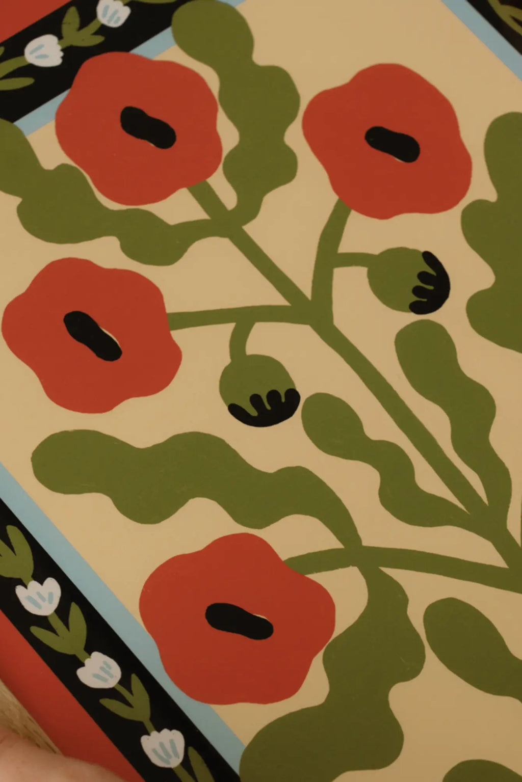 The Poster Club Red Poppies, a vibrant graphic botanical art print featuring bold red poppies with black centers, olive green leaves, and stems on a light beige background. The modern folk art style is accented by a black and light blue border with small white and green floral motifs. Perfect as Scandinavian wall art for minimalist living rooms, bedrooms, or a colorful addition to a contemporary gallery wall.