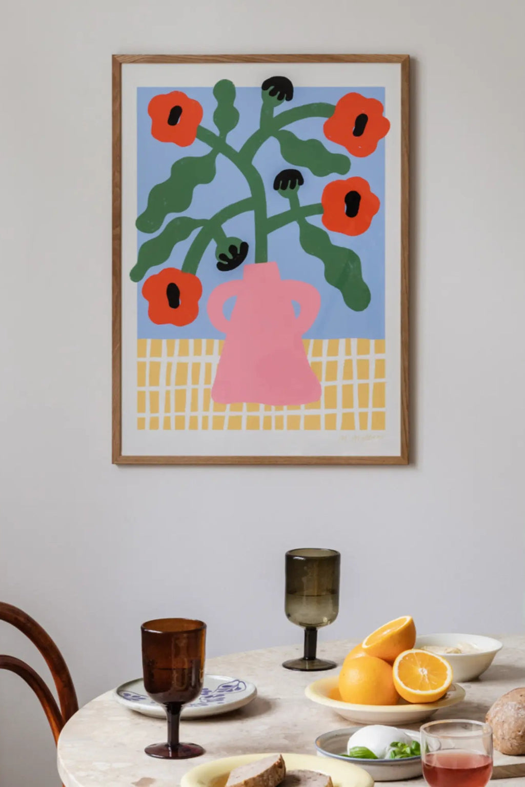 The Poster Club Red Poppies in Pink Vase, a vibrant abstract floral art print featuring bold red poppies with black centers, green organic stems and leaves, set in a minimalist pink vase against a light blue background, grounded by a yellow grid pattern. This contemporary wall art comes framed in natural oak, perfect for adding a pop of color to modern dining rooms, Scandinavian living rooms, or minimalist bedrooms.