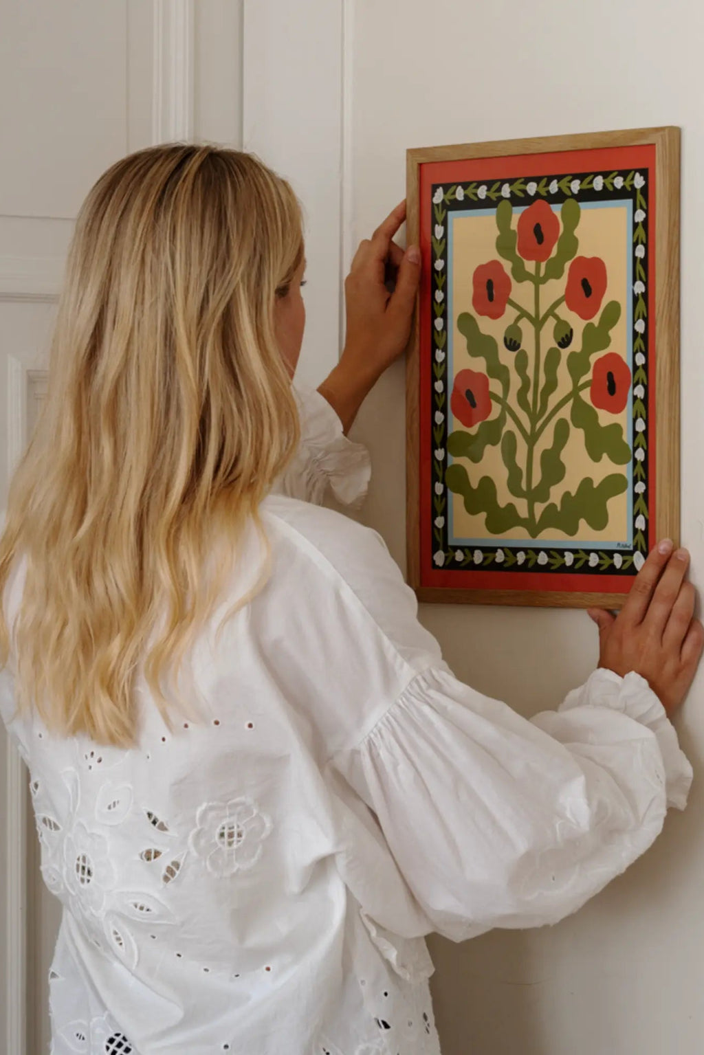 The Poster Club Red Poppies, a vibrant folk art print featuring stylized red poppies and dark green leaves on a light cream background, framed by a layered border in terracotta red, black with white floral accents, and a thin light blue edge. This colorful botanical wall art comes in a natural oak frame, ideal for adding rustic charm to Scandinavian living rooms or a contemporary gallery wall.