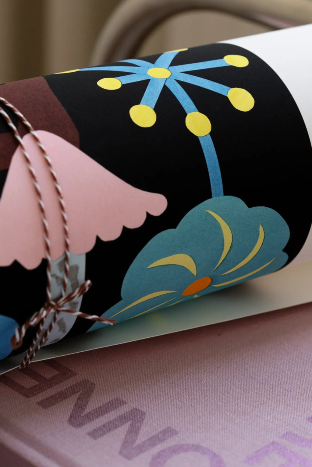 The Poster Club The Garden of the Night, a vibrant abstract floral poster featuring bold graphic shapes in light blue, yellow, orange, pink, and brown against a striking black background, rolled and tied with brown and white twine. This modern art print is perfect for adding colorful, playful wall art to a contemporary living room, bedroom, or nursery.